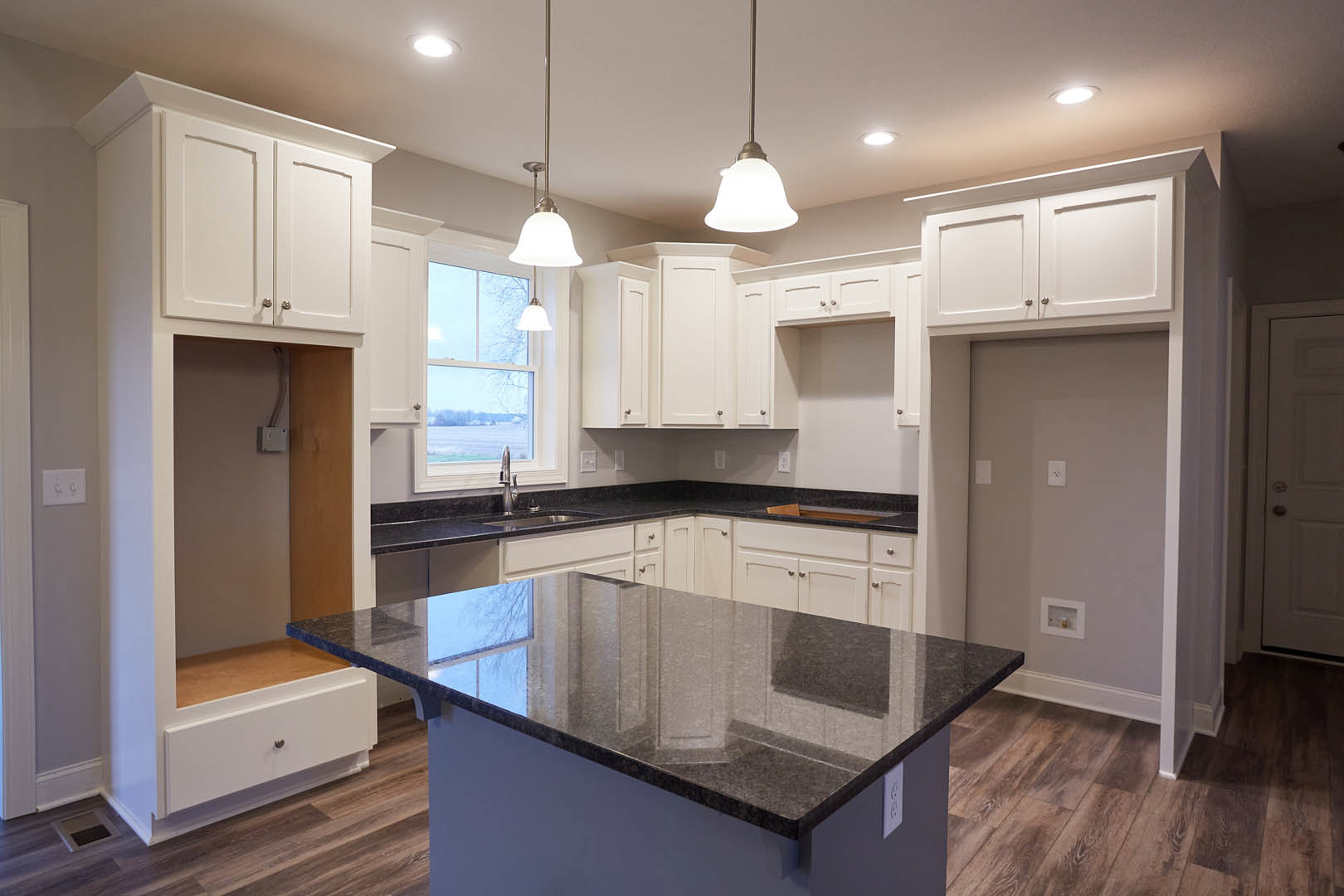 Modern kitchen featuring black stone countertop, white cabinetry, stainless steel sink, silver door handle, ceiling light fixture, large window overlooking snowy landscape, and