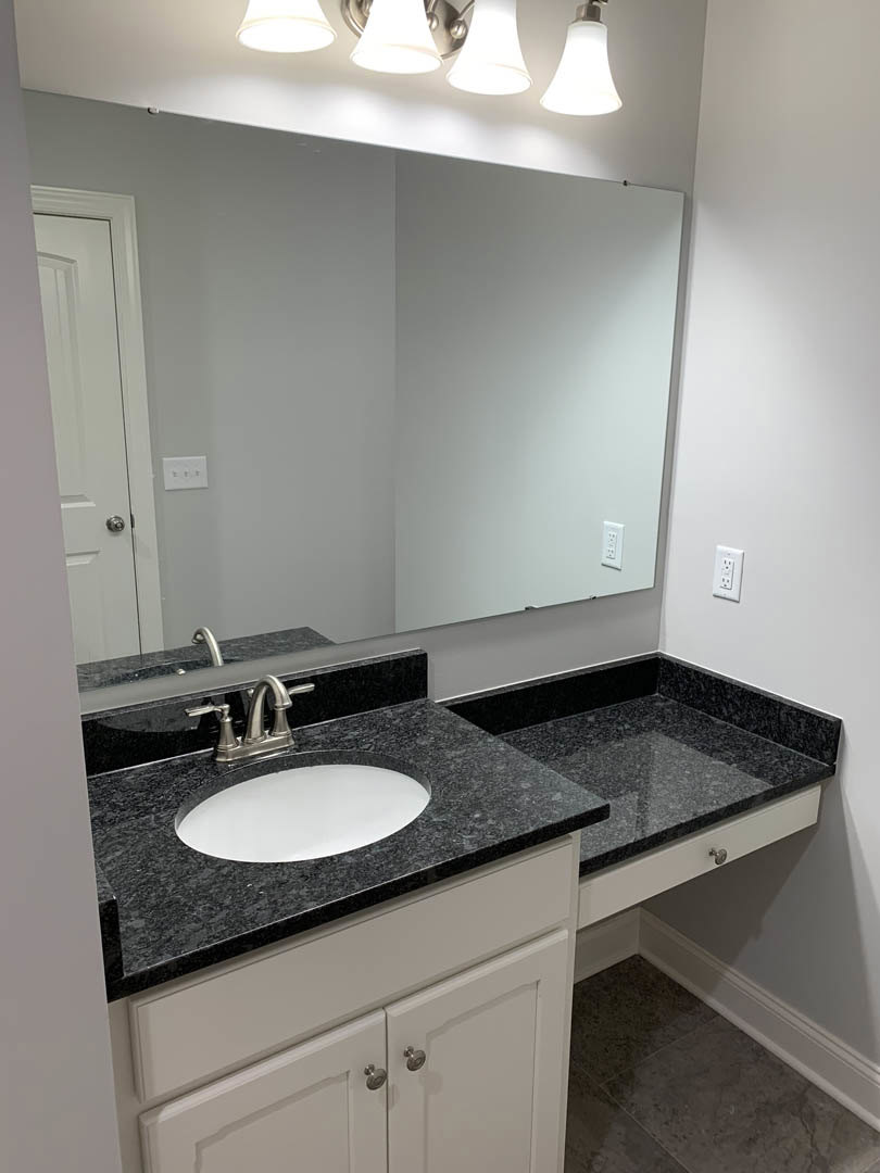 White porcelain sink with chrome faucet set in a stone countertop, rectangular wall mirror above, light fixture reflected, gray tile flooring, white cabinetry visible below.