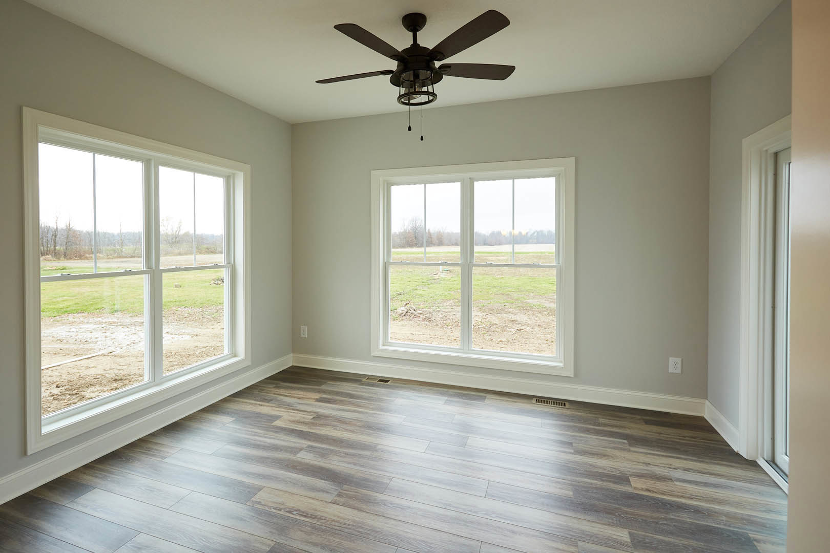 Wood flooring in a bright room with large windows overlooking grass, ceiling fan with light fixture centered above.
