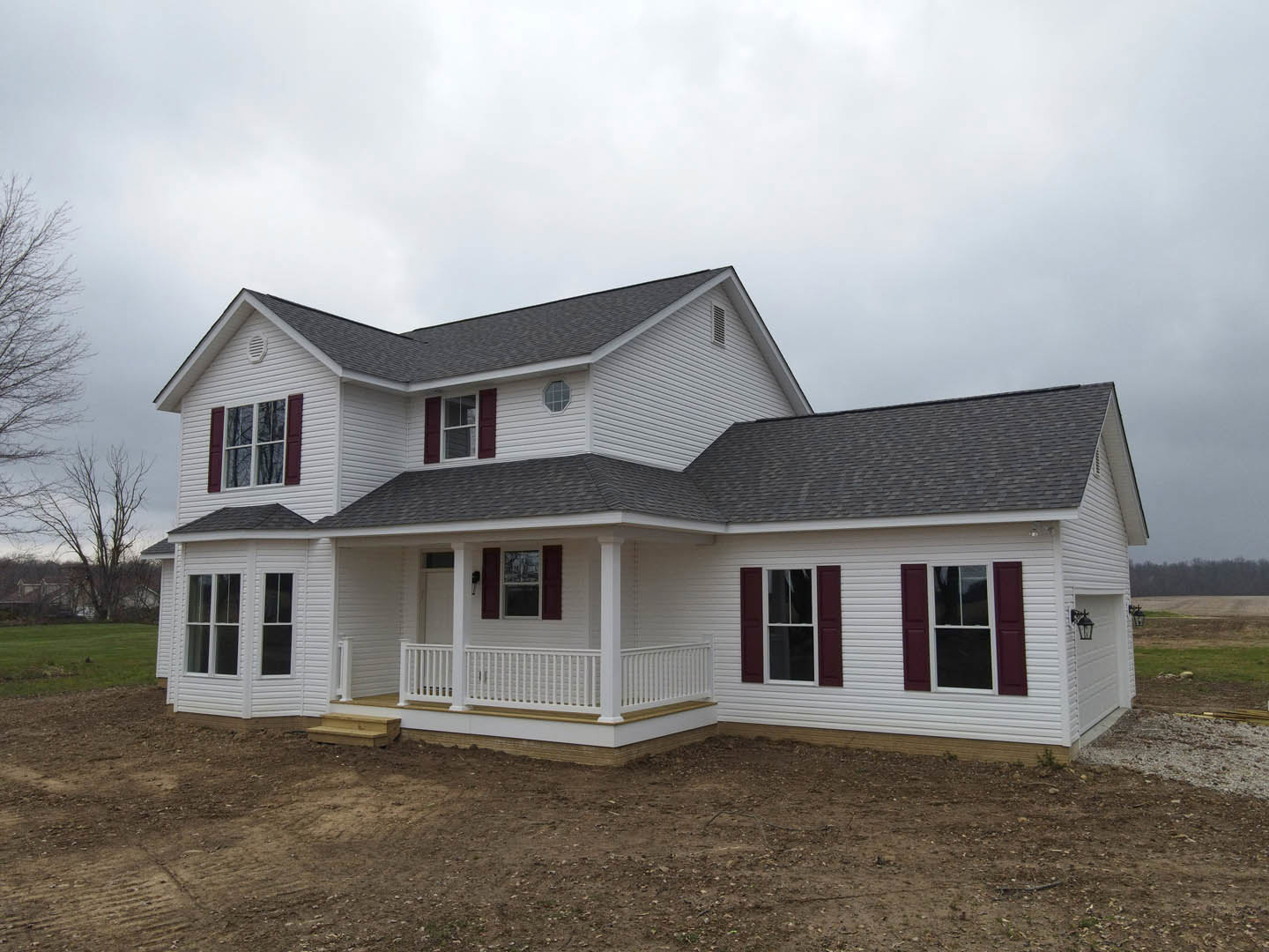 White house with red shutters and white-framed windows, porch with white railing, dirt yard in foreground, tree branches and cloudy sky in background