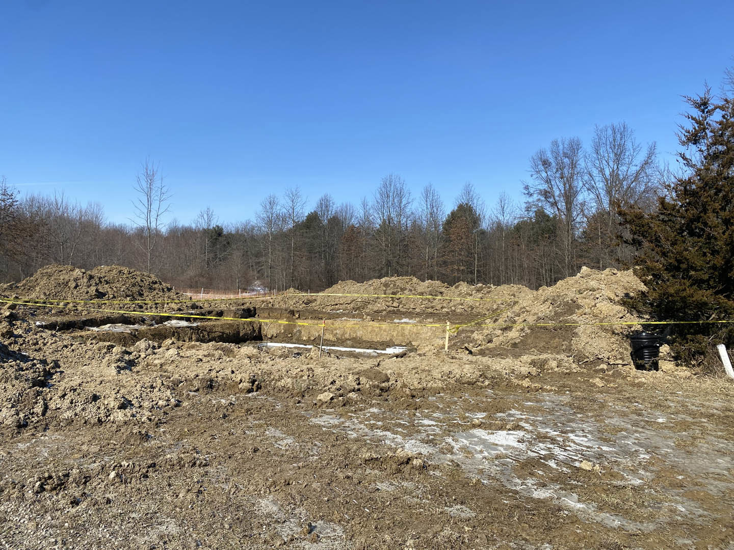 Dirt field bordered by leafless trees under a clear blue sky, yellow tape marking the ground, black bucket placed on soil