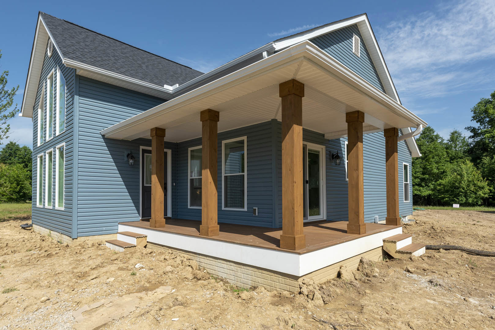 Two covered porches with white pillars and wooden posts, black shutters on windows with blinds, light siding, tree branches with green leaves in foreground