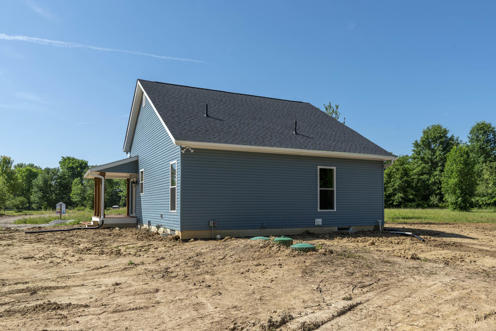 Partially built house with exposed framing, white window frame, dirt lot scattered with green containers, trees and another building in the background, shingled roof visible