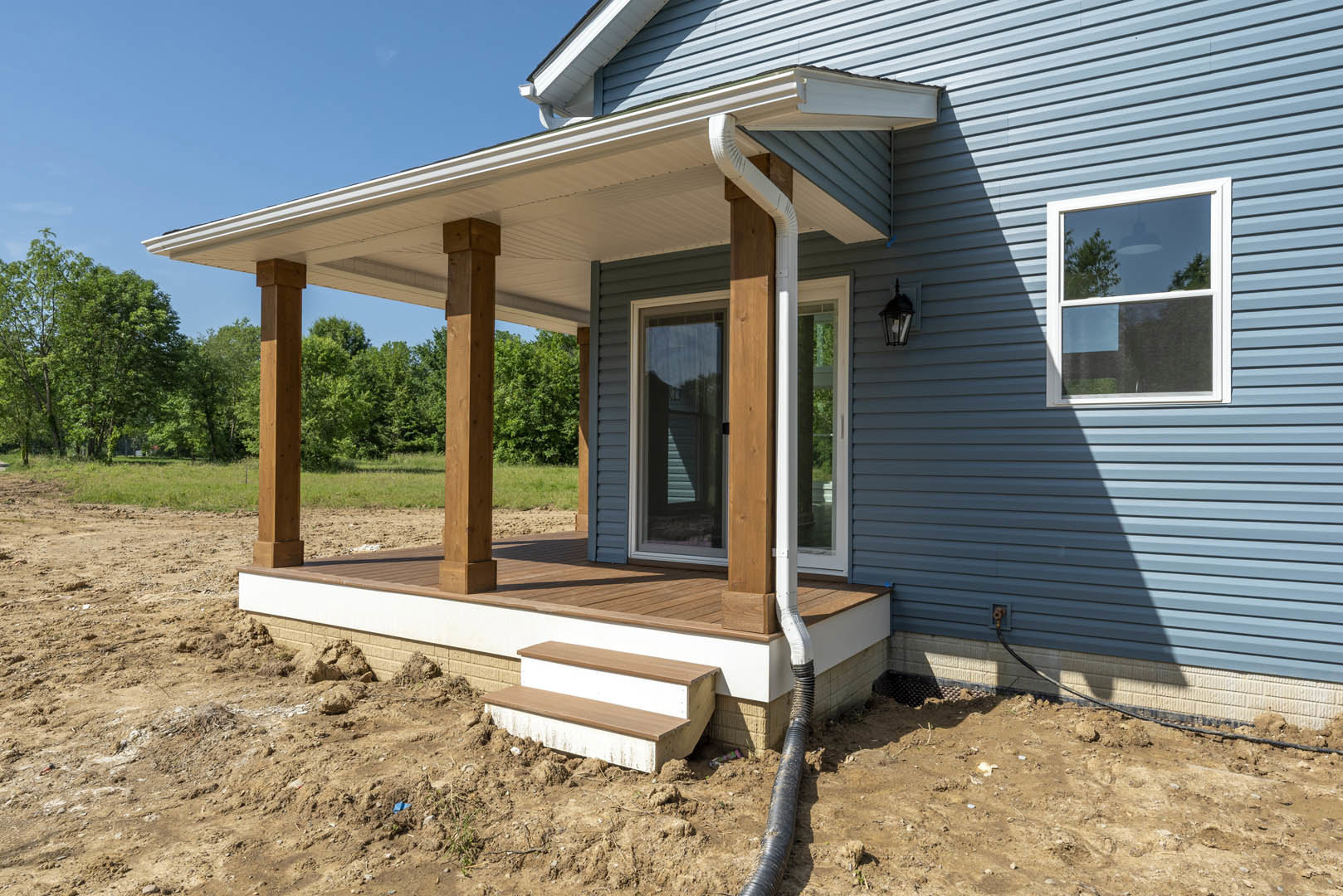 Two-story home with gray siding, white trim, covered front porch, wood door, white-framed windows with blinds, wall-mounted light fixture, concrete steps, landscaped yard with