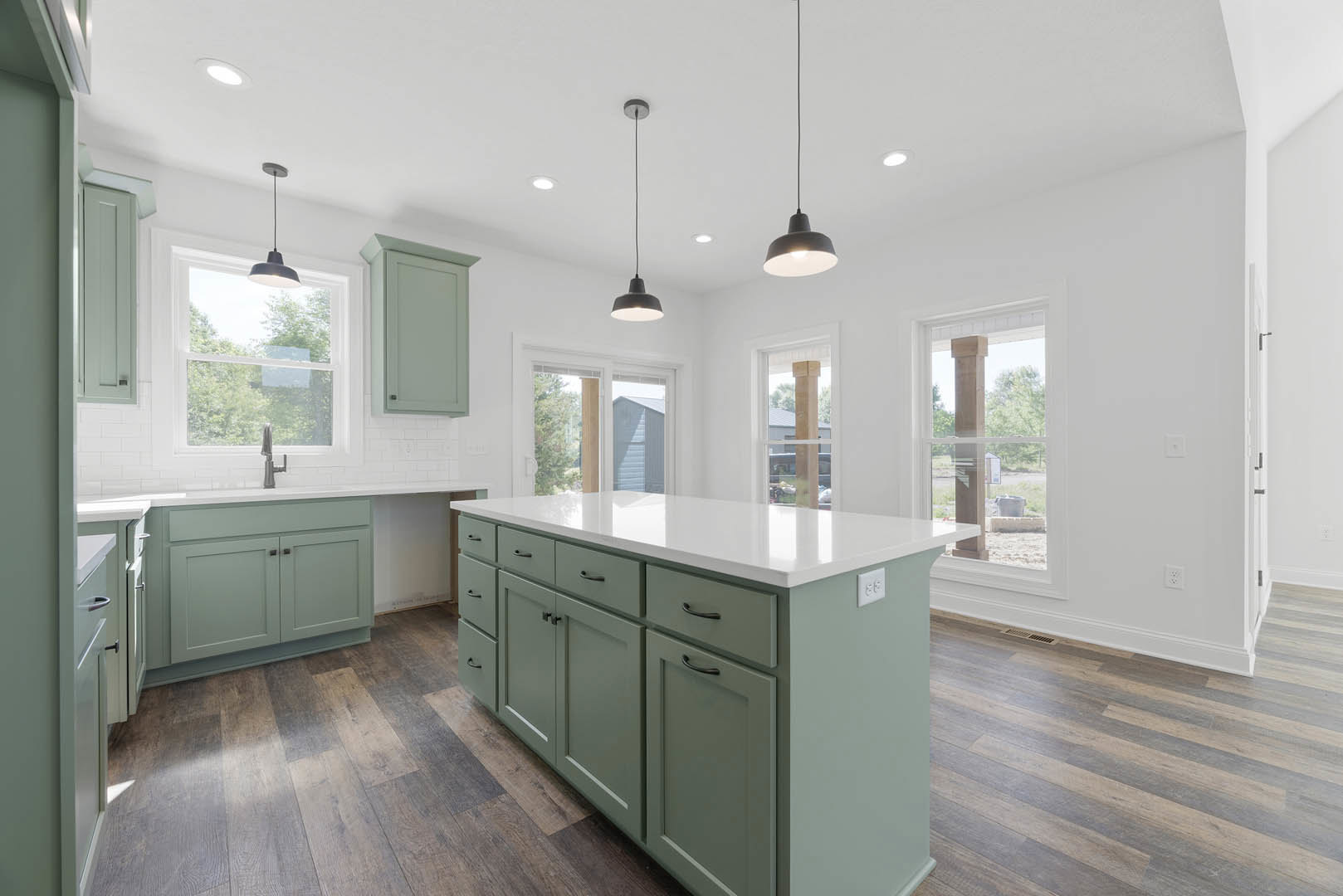 Kitchen with green cabinets, wood flooring, white countertop island with drawers, white door with white frame, black wire along wall, pole with flag.