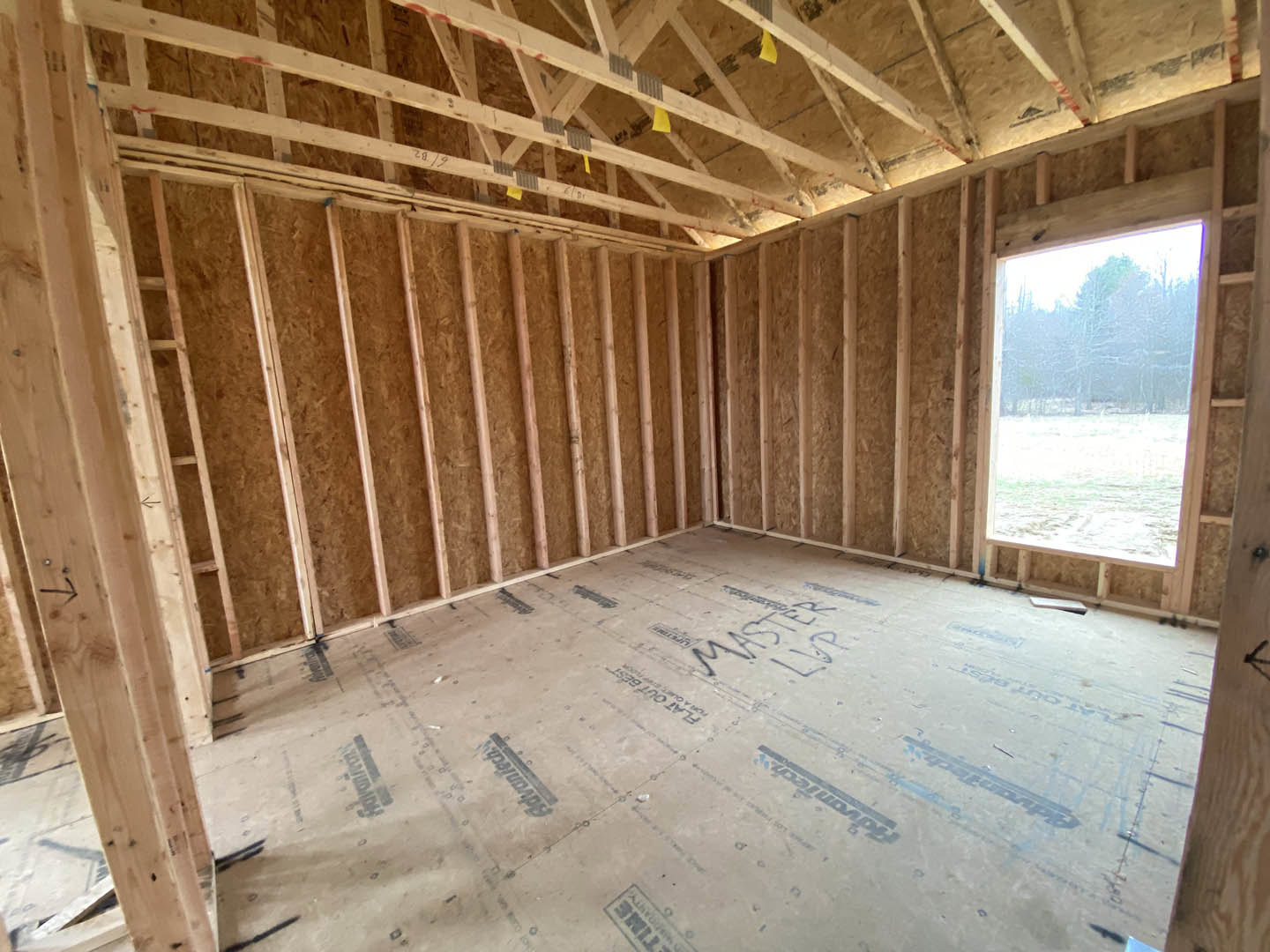 Exposed wood ceiling beams, unfinished subfloor with construction markings, large window overlooking forested landscape, yellow insulation visible between roof trusses