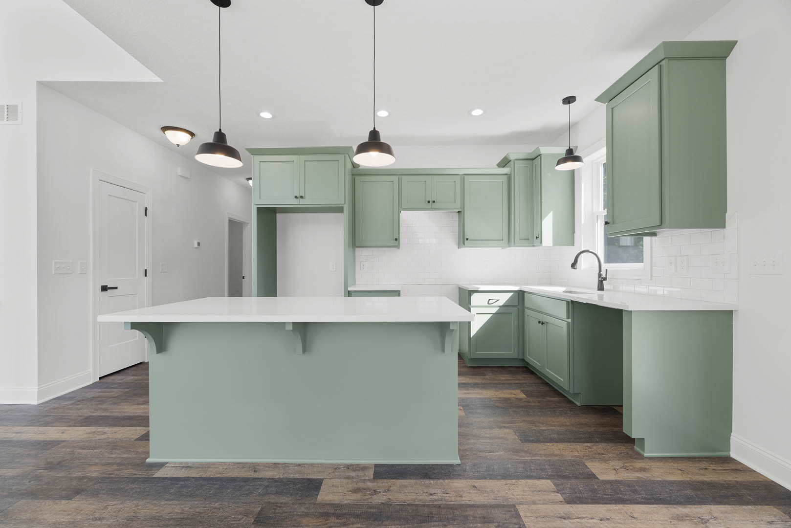 Green kitchen cabinets with white quartz countertops, black hardware, white tile backsplash, pendant lights with black cords, and light wood flooring.
