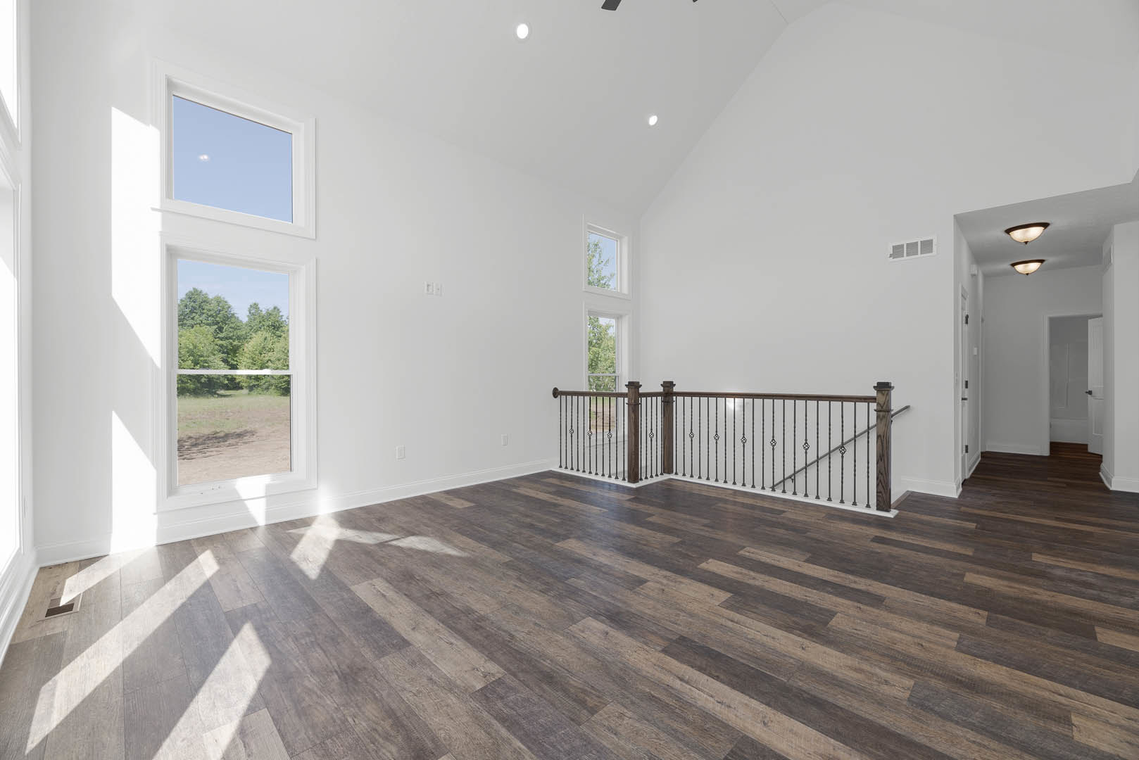 Open-concept room with wide plank wood flooring, metal railing along staircase, large windows overlooking trees, white walls, and modern finishes