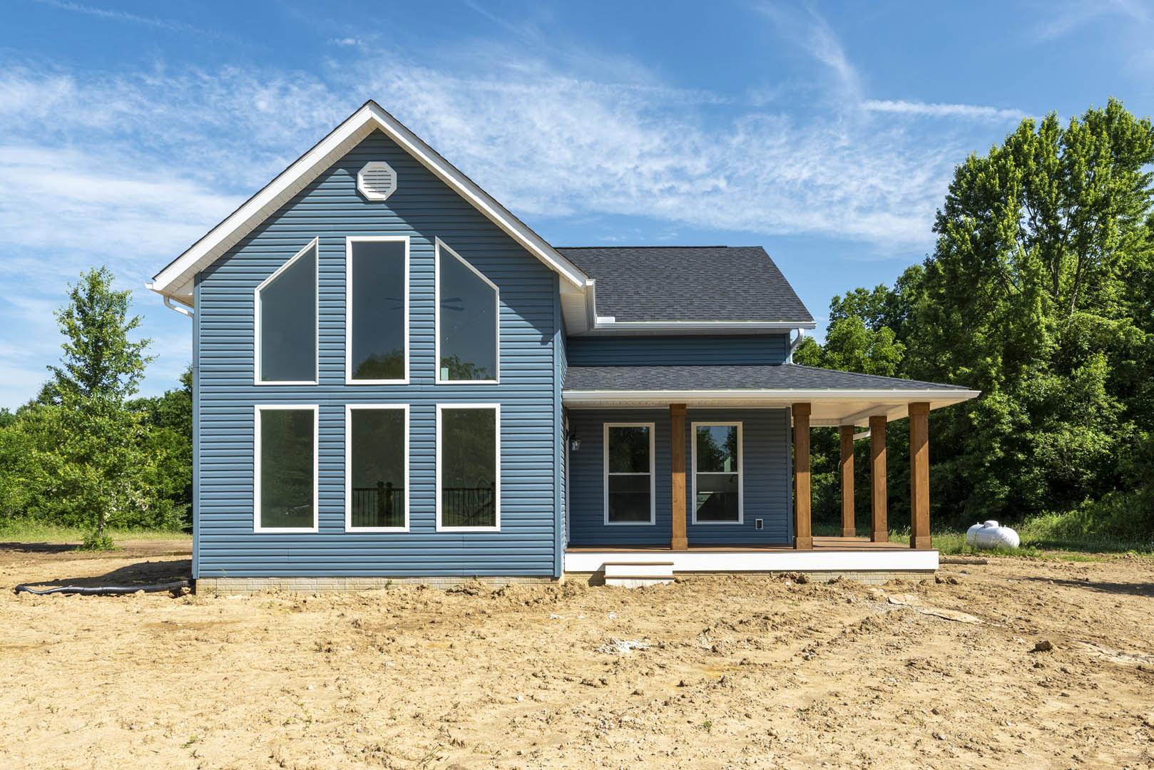 Blue house under construction with white-trimmed windows, blue siding, white vent, dirt ground, and trees in the background