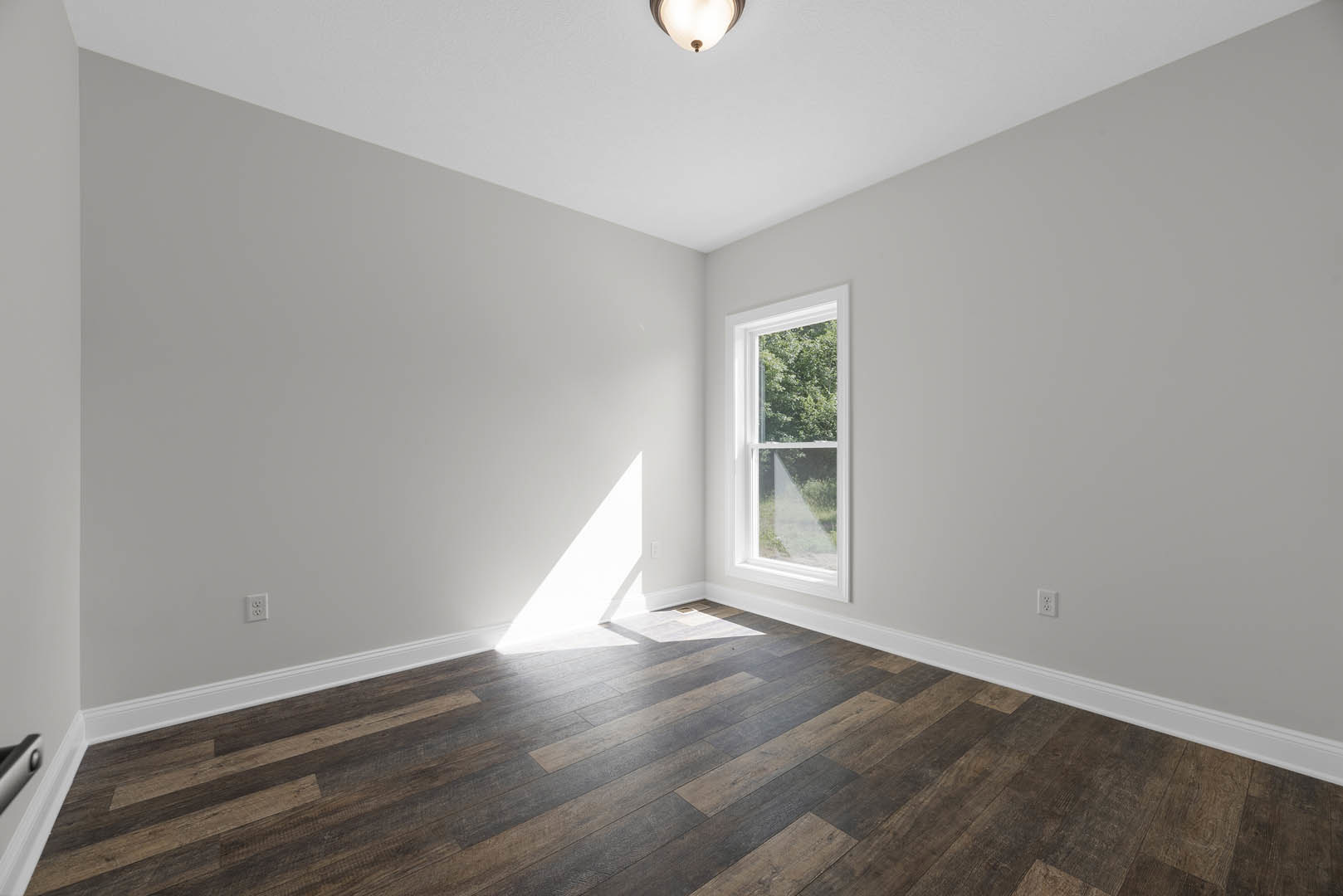 Sunlit room featuring wide-plank hardwood flooring, white-framed window overlooking trees, smooth plaster walls, and recessed ceiling light.