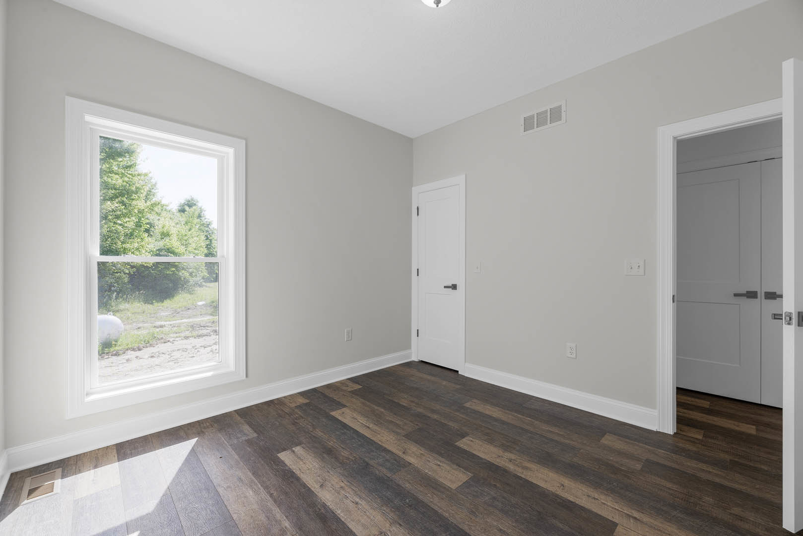 Wood flooring with white baseboard trim, white door featuring a black handle, large window framing leafy trees outside, and minimalist white walls