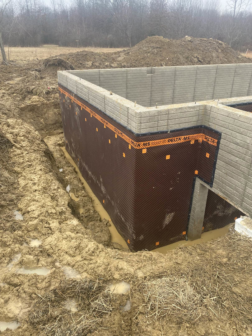 Concrete foundation with exposed rebar and large excavation pit, surrounded by dirt, construction materials, and trees in the background