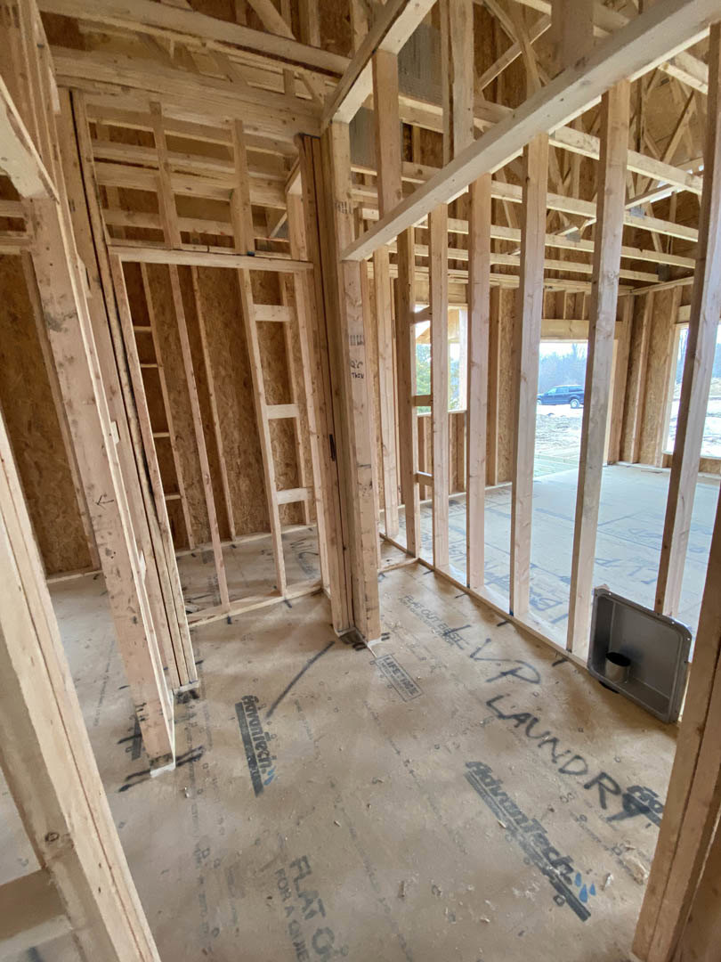 Wood framing and beams inside a partially constructed house, exposed lumber walls, metal bin embedded in framing, construction sign and logo visible, car parked at site.