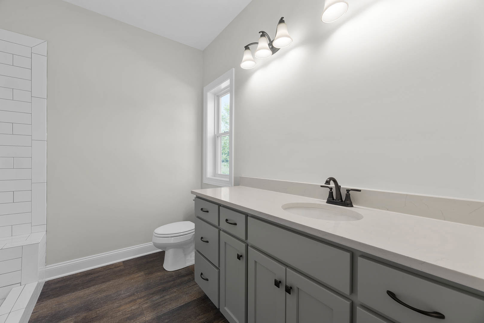 White bathroom with black faucet on white countertop, white toilet, white brick accent wall, three-light fixture, window close-up, and dark cabinetry drawers.