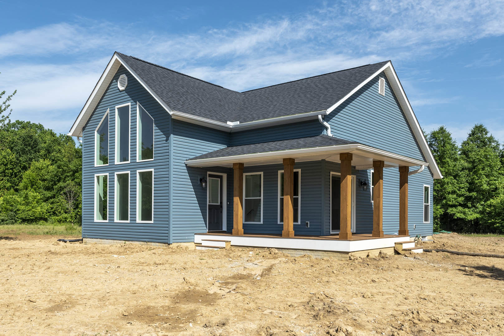 Partially built house with exposed wooden framing, white window frames, covered porch, dirt yard, and blue sky; trees visible in background