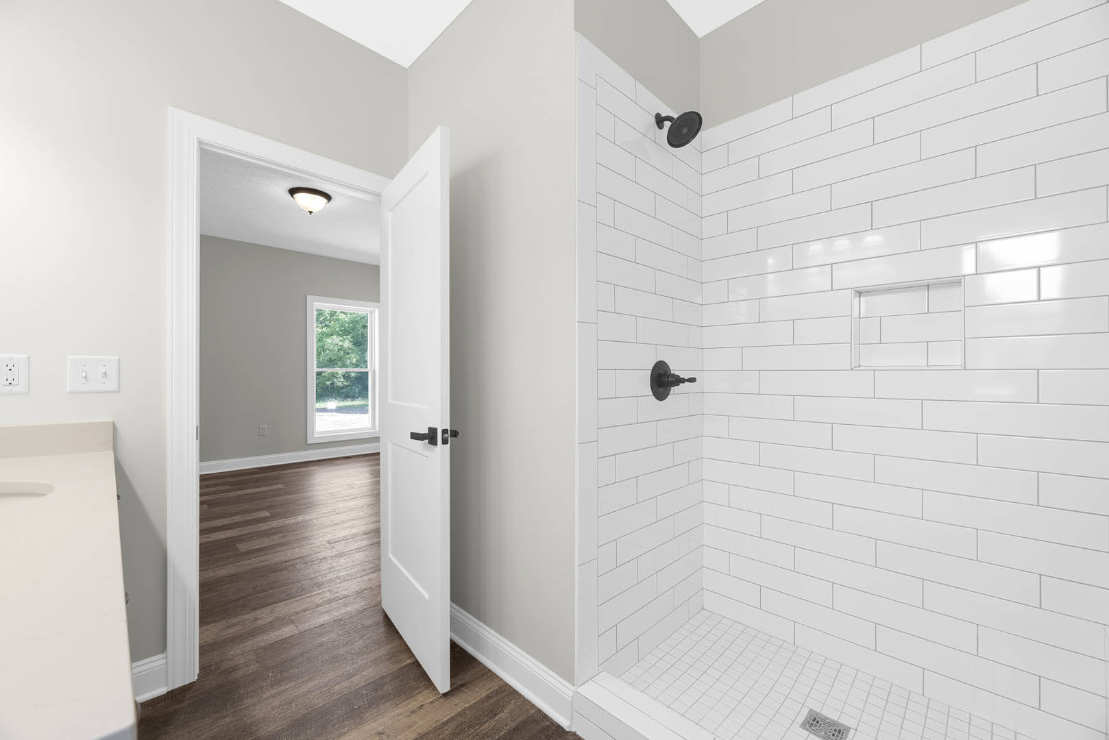 Bathroom with wood flooring, glass-enclosed shower featuring black shower head, white walls, window overlooking trees, white countertop with black round accessory, double white
