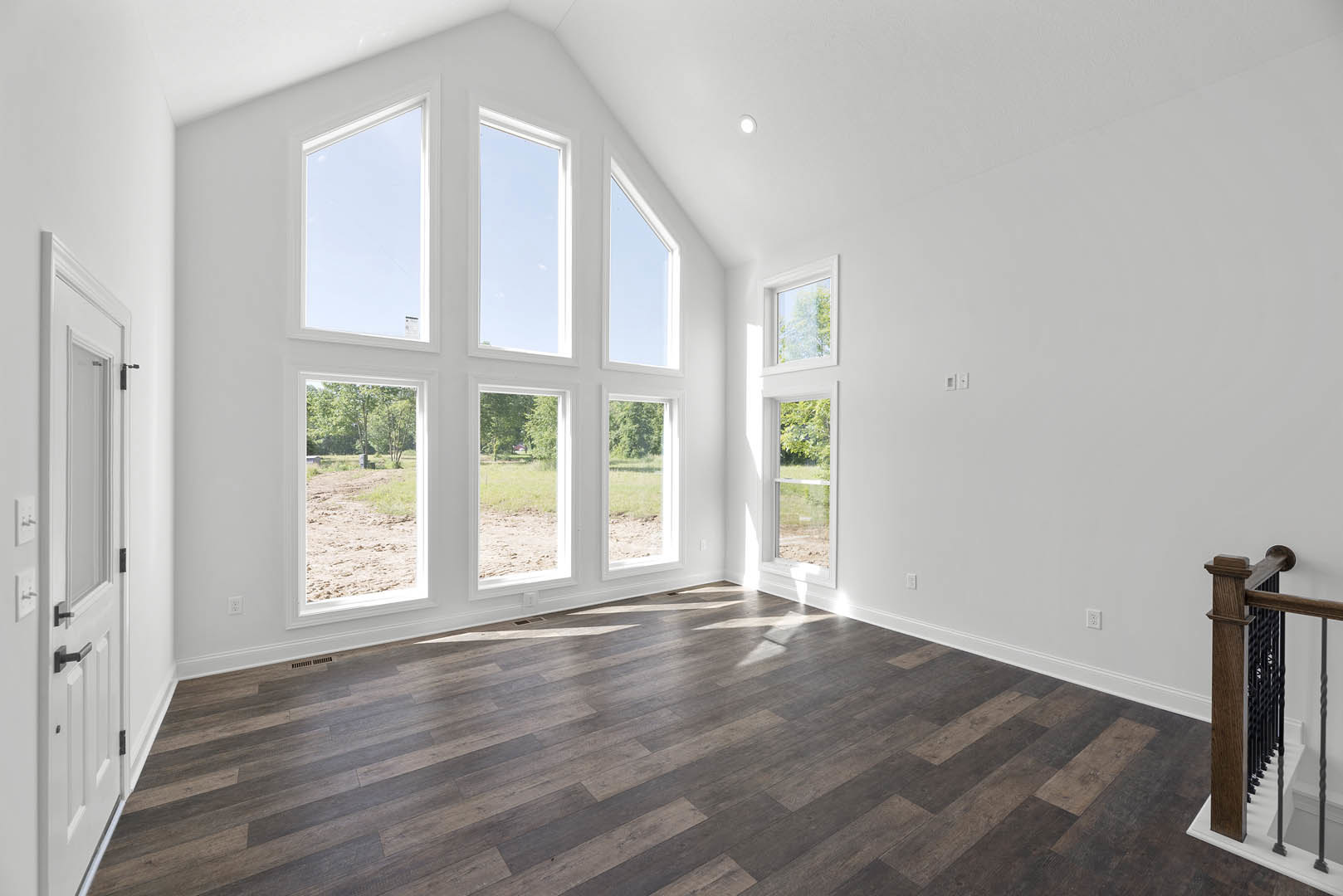 Spacious room featuring wide plank wood flooring, multiple tall windows framed in white, and a modern metal railing adjacent to a crisp white wall with blue sky visible through the