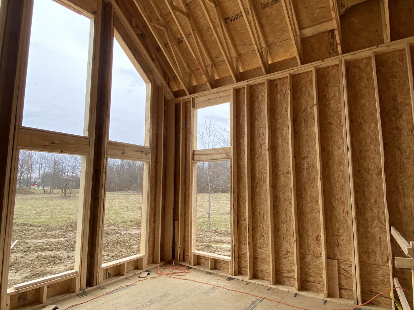 Room under construction with exposed wood framing, large windows, and natural light; grassy field and trees visible outside