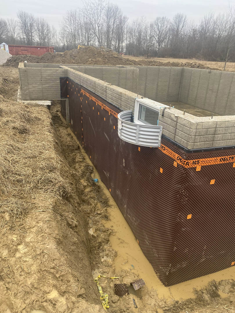 Concrete foundation with exposed soil, white framed window set in a partial wall, grassy field bordered by trees, red construction container with white lettering