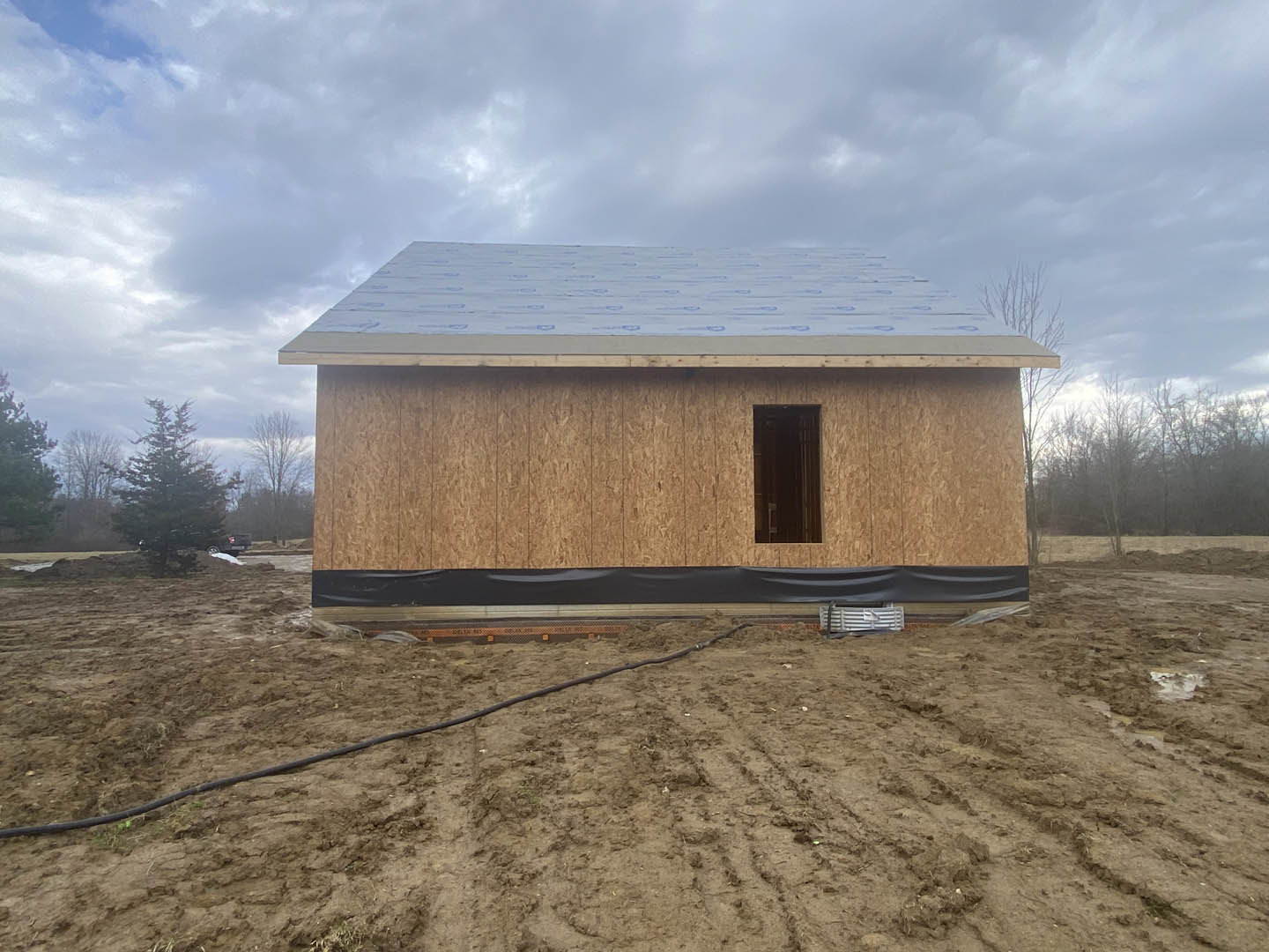 Partially built house with exposed framing, unfinished door opening, dirt lot, black hose on ground, and cloudy sky overhead
