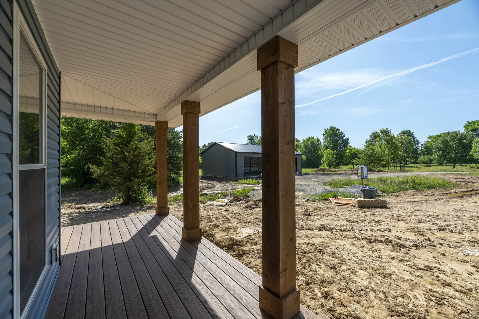 Wooden porch with white roof, sturdy pillars, and adjacent grey shed with open door; window and deck visible, surrounded by trees and plants.