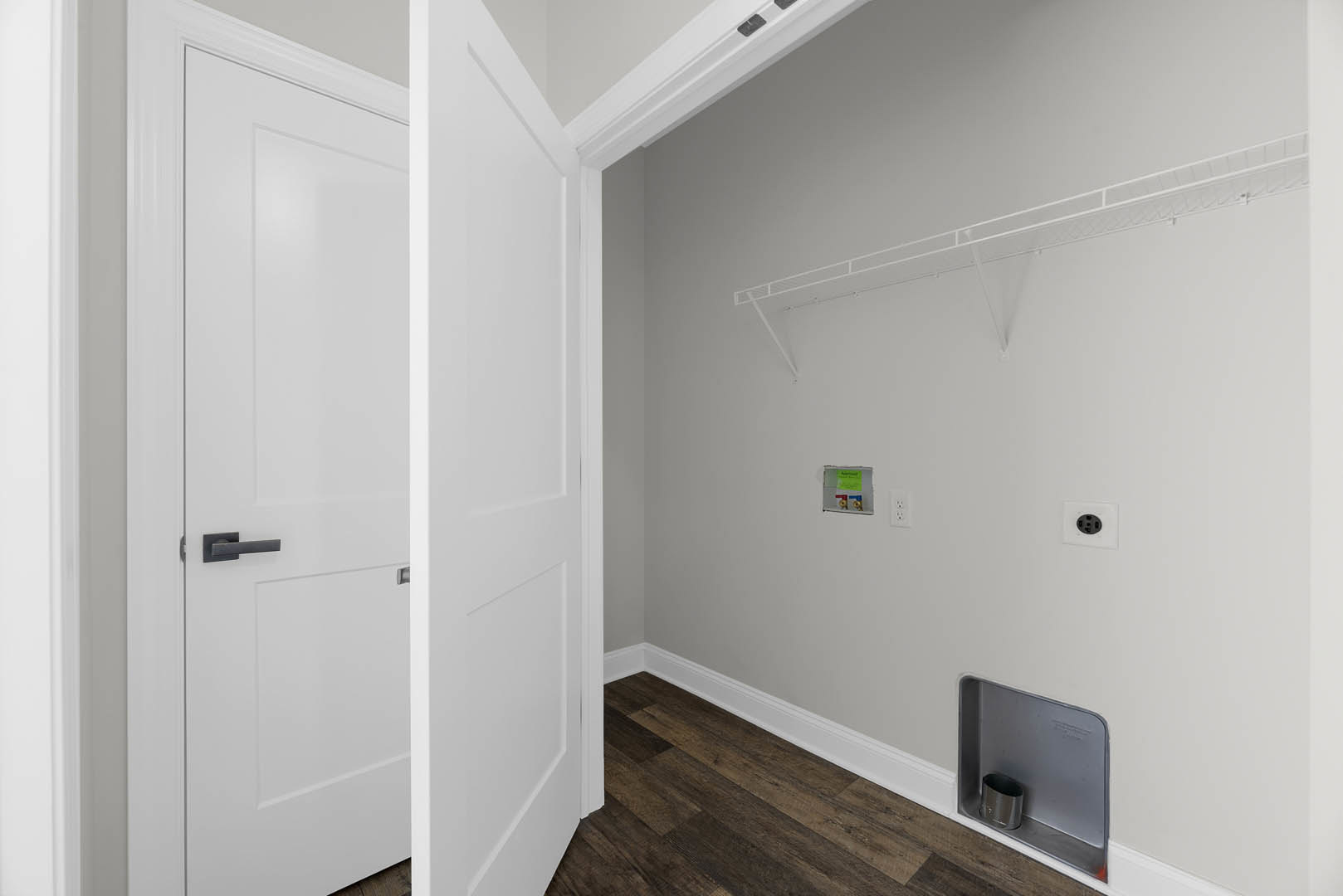 White door with silver handle open to a room with light plaster walls, black electrical outlet, and wood flooring; silver trash can with black lid visible near entry.
