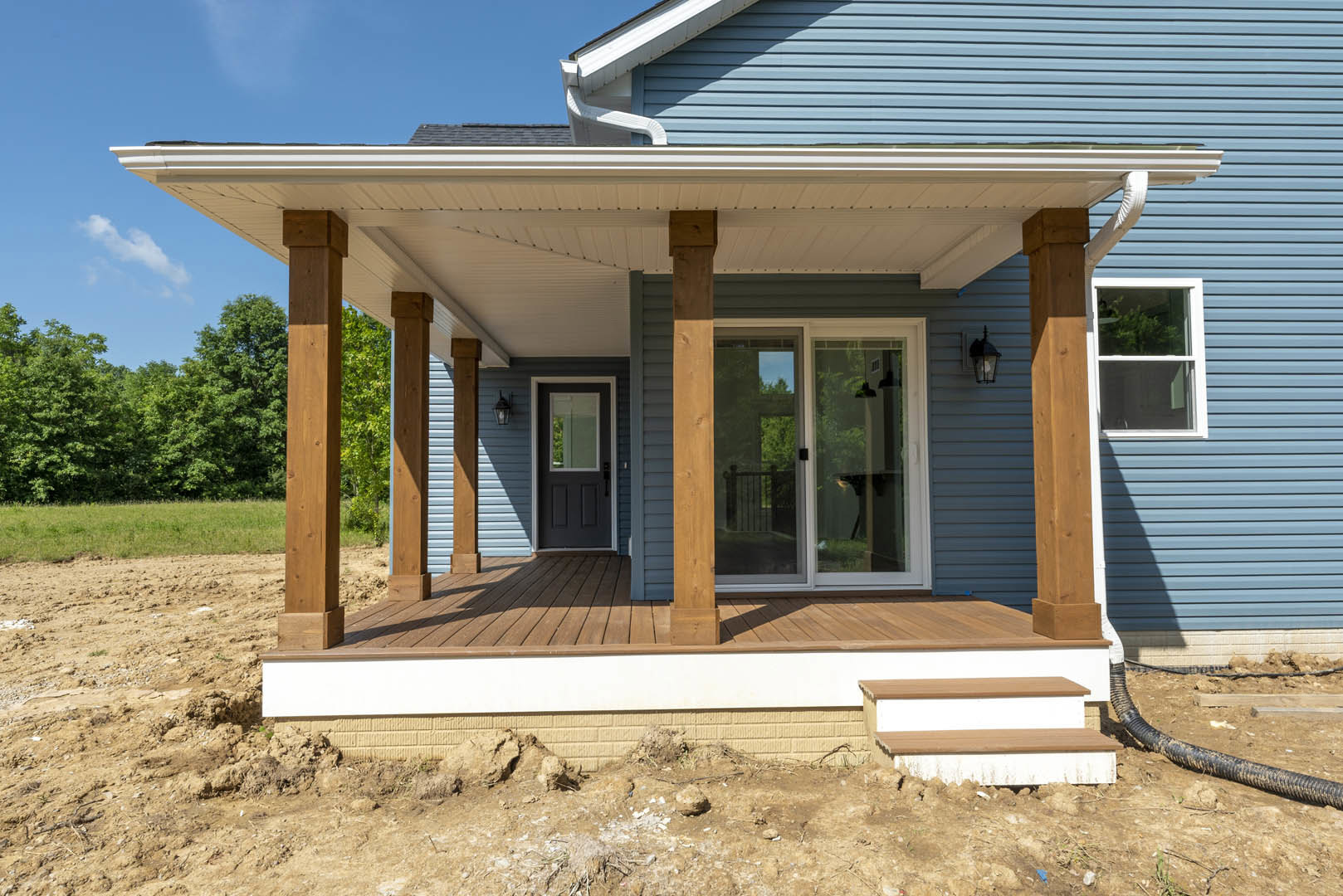 Two porches with white railings and columns, glass door with wooden frame, white-trimmed windows, gray siding, small shed in background, green tree and fence visible