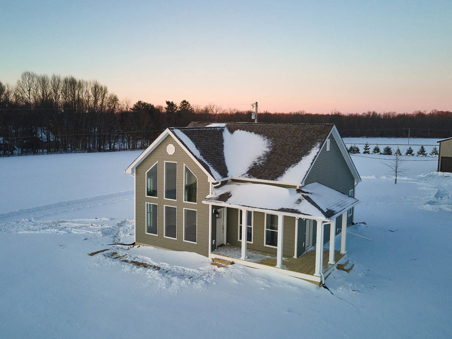 Snow-covered roof and eaves of a residential home with light siding, surrounded by bare trees under a blue sky with scattered clouds