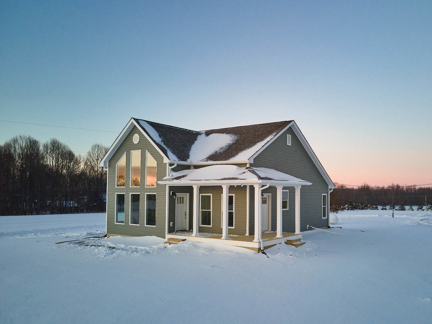 Two-story cottage with snow-covered roof and porch, surrounded by leafless trees, white door with glass panes, large windows, and snowy yard.