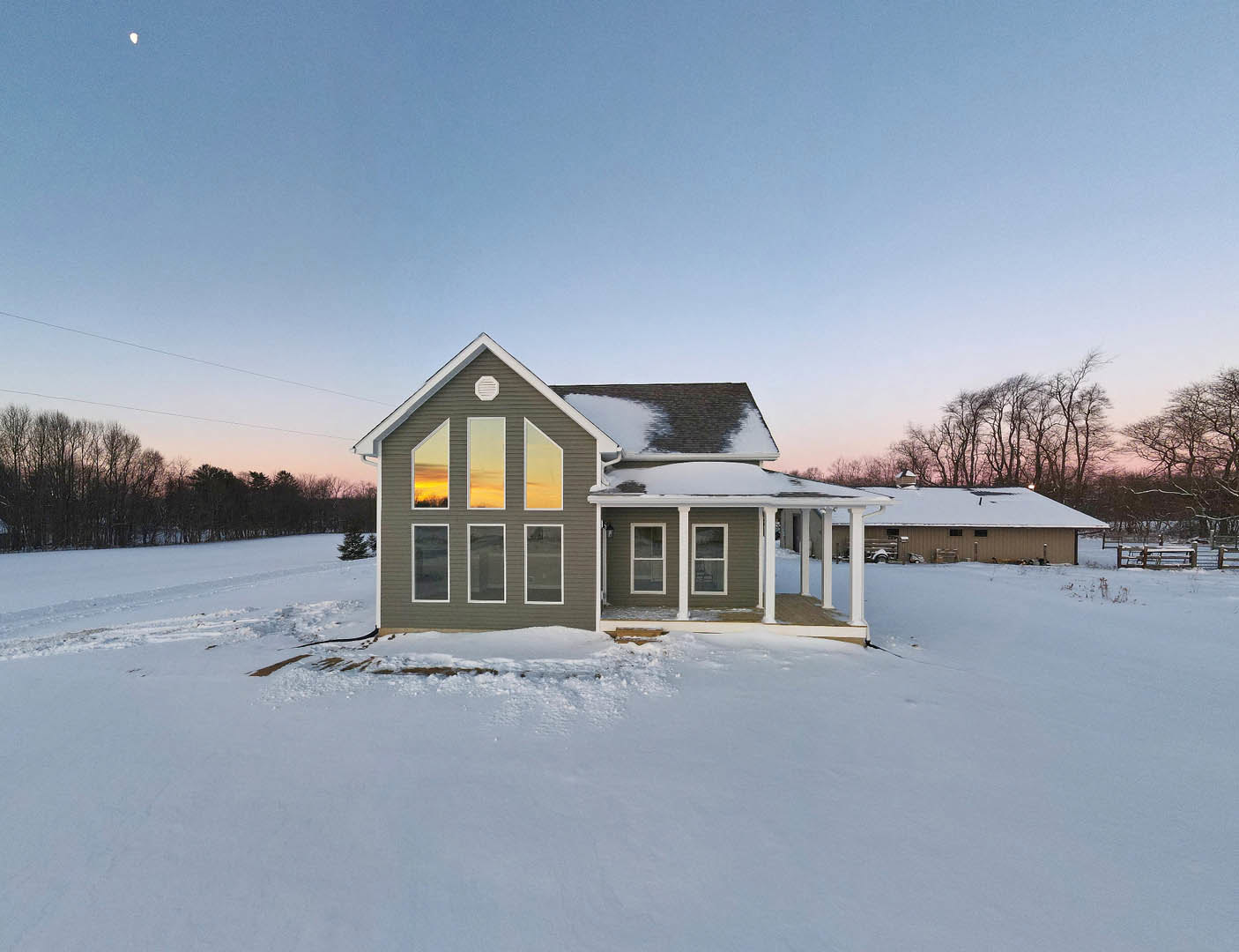 Two-story home with large windows, snow-covered roof and ground, surrounded by leafless trees under a cloudy winter sky