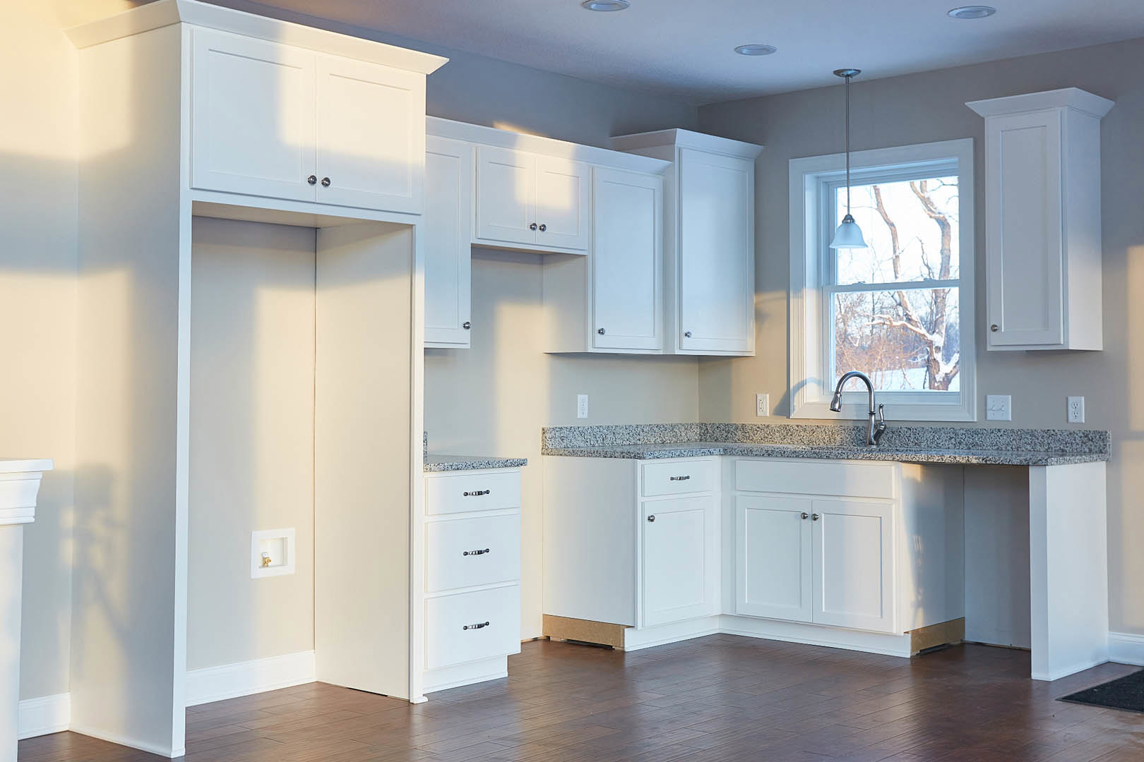 White kitchen with granite countertops, sunlight streaming through window, stainless steel faucet, wooden floor with white baseboard, white door and drawer fronts