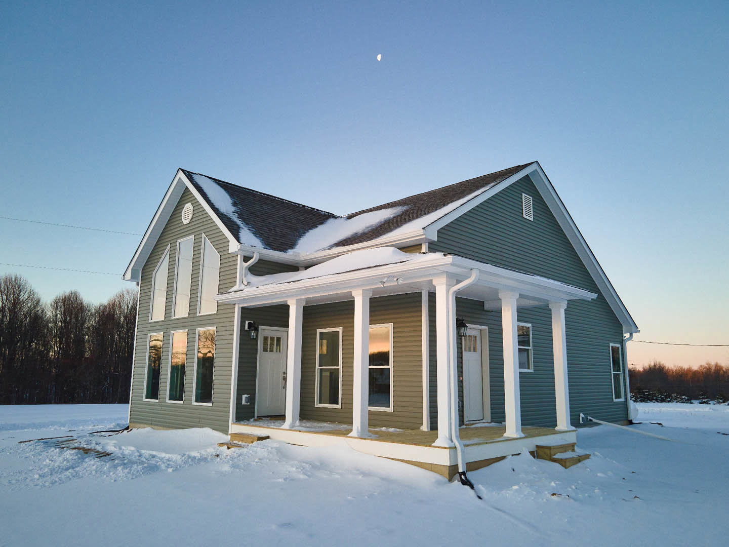 Two-story house with white siding, black trim, and snow covering the roof and ground; white-framed windows and a white front door with black hardware; leafless trees in the snowy