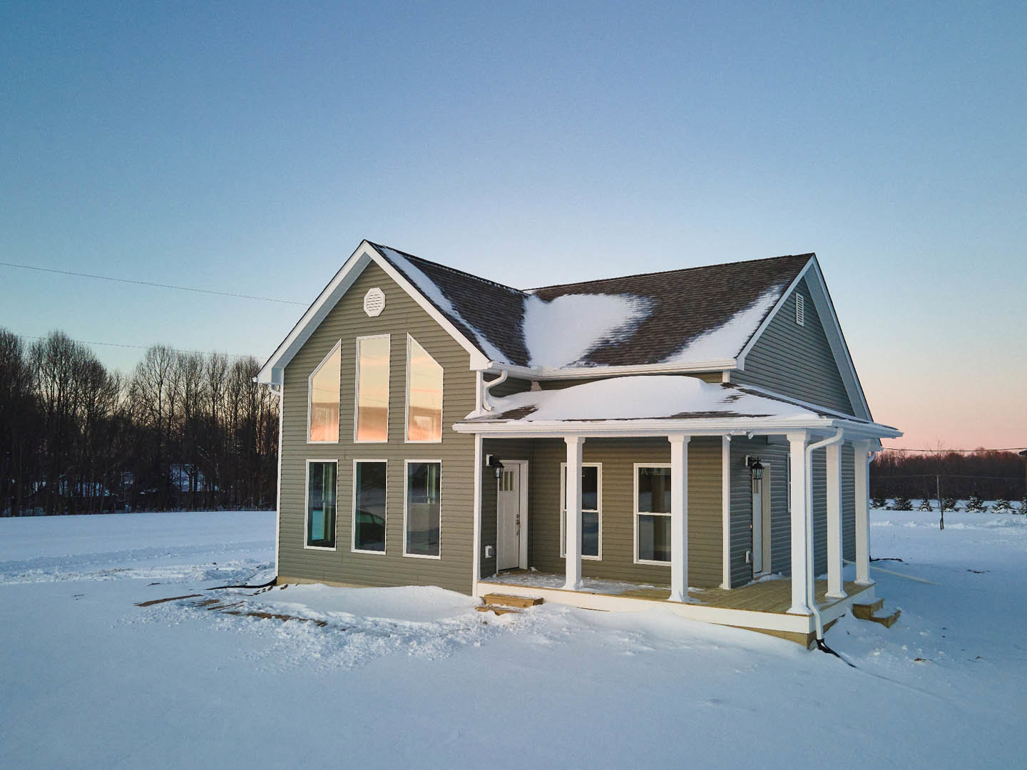Modern white house with metal-framed windows, snow-covered roof and yard, surrounded by leafless trees under a pale winter sky