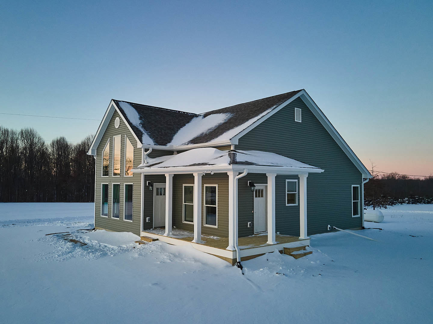 Two-story home with white-framed windows, snow-covered roof and yard, surrounded by leafless trees under a pale winter sky