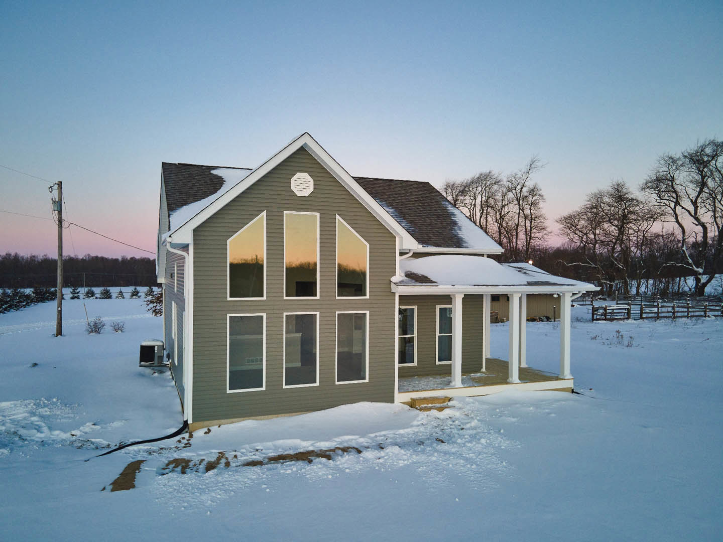 Two-story house with white siding and white-framed windows, snow covering the ground and roof, leafless trees in the background, white vent visible on exterior wall