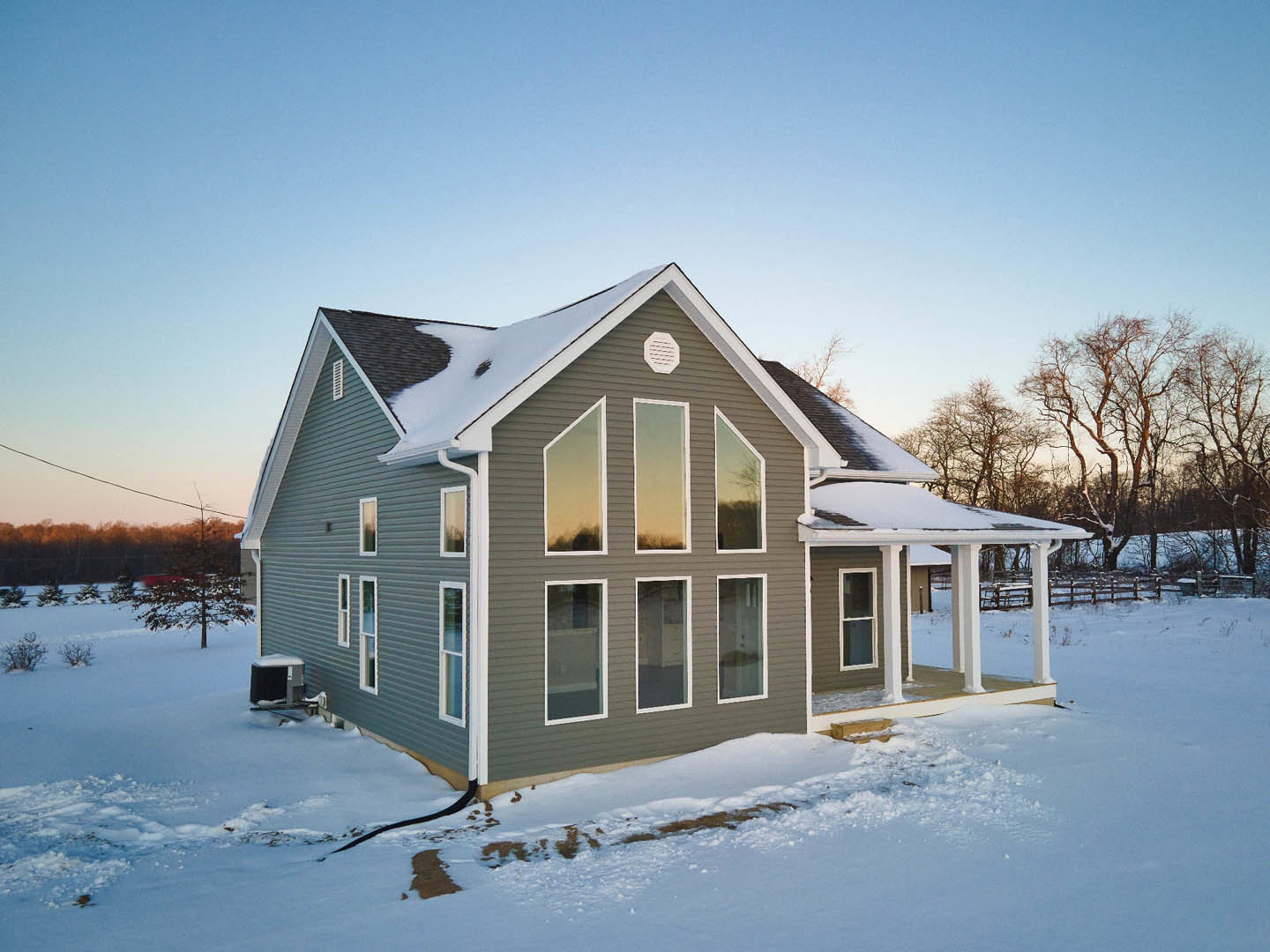 Modern cottage with white exterior walls, large windows, and snow-covered ground; leafless trees and pale yellow sky in the background.