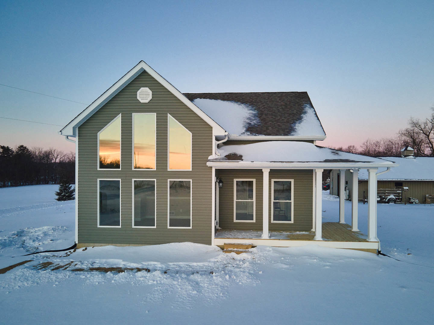 Two-story cottage-style home with multiple white-framed windows, snow covering the ground and roof, sunset sky reflected in glass, bare trees in background, white vent visible on