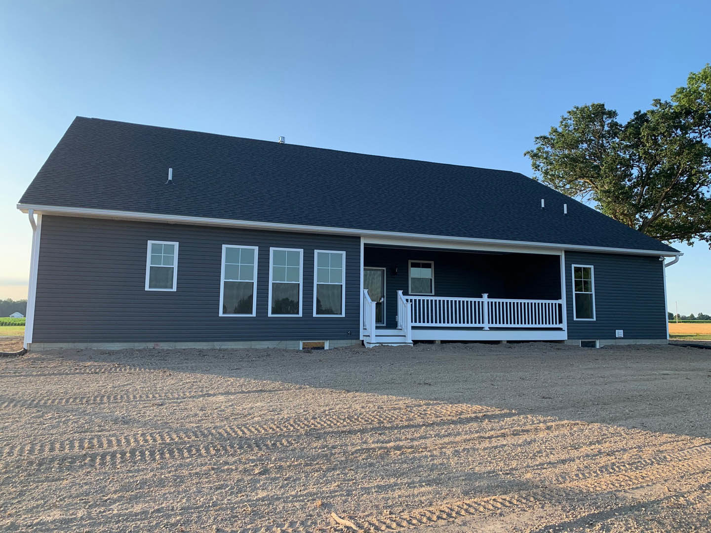 Two-story house with white porch railing, dirt road and tire tracks in front, white-framed windows, gray siding, shingled roof, tree in background.