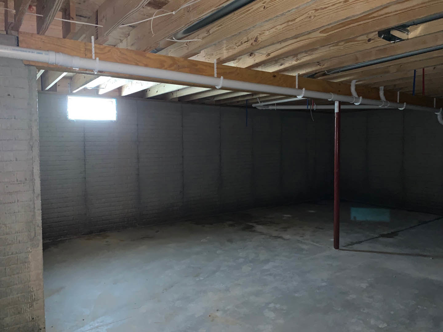 Exposed brick wall and wooden beam ceiling in a basement room with white flooring, visible pipes, and a window.