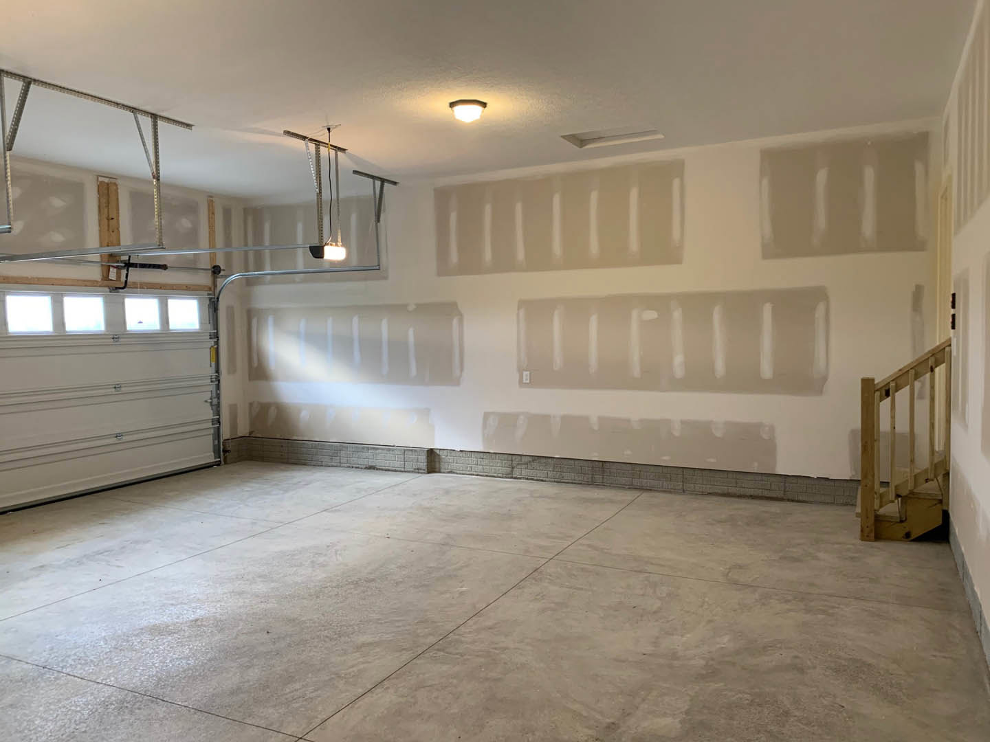 Concrete-floored garage with white plaster walls, wooden railing along stairs, square window letting in natural light, ceiling-mounted light fixture, white garage door with subtle