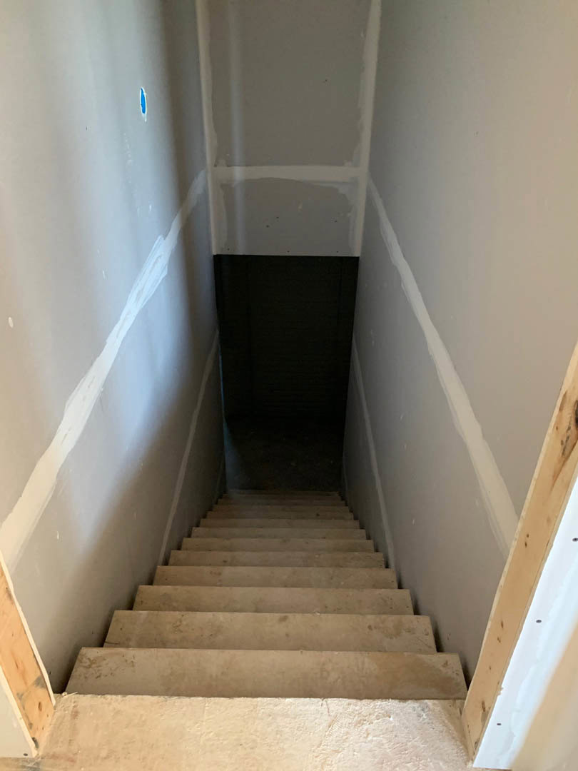 Wood staircase with white risers ascending beside a smooth white plaster wall, leading to an upper room with light-colored flooring