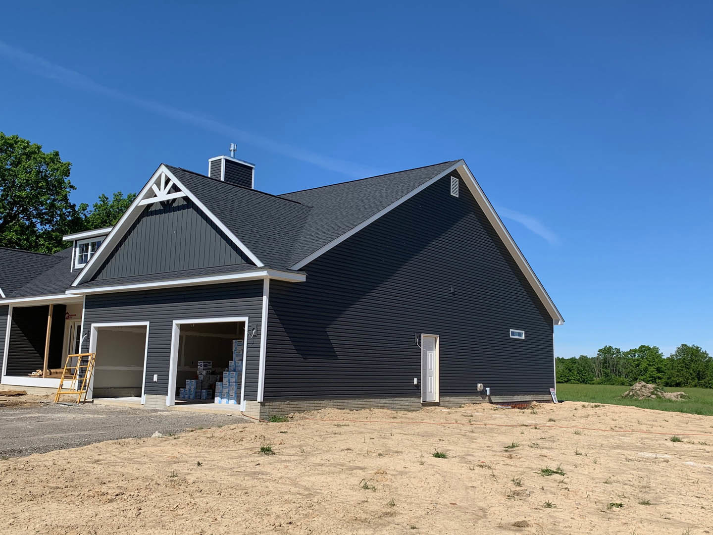 Modern home with attached garage, white exterior walls, yellow ladder leaning against garage wall, stack of boxes inside garage, dirt driveway bordered by red rope, trees and blue