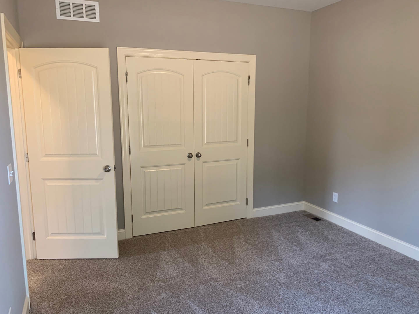 Carpeted room featuring a white double door with silver knobs, a single white door with a silver knob, wall vent, and a white square cabinet with a black handle