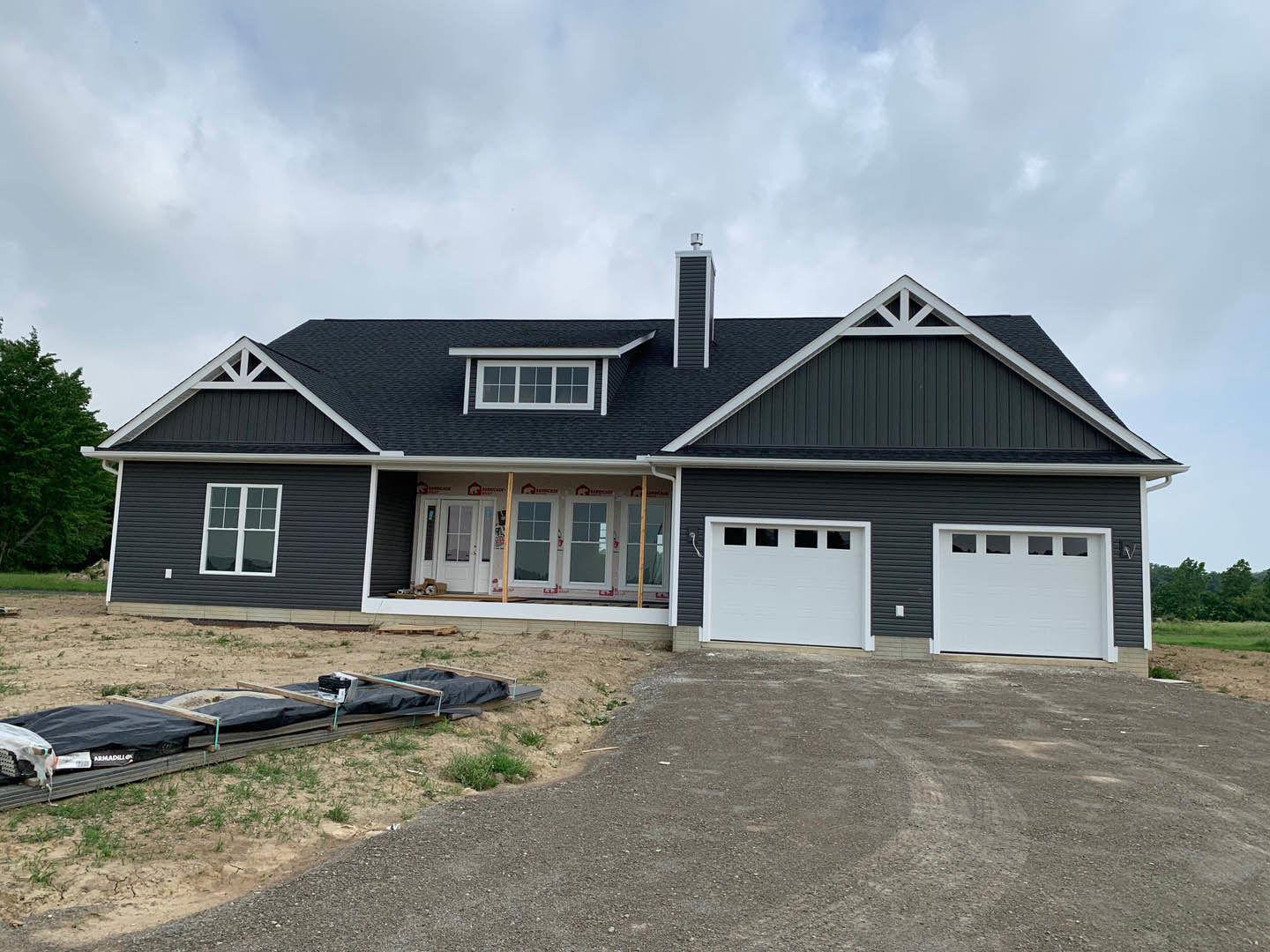 Two-story custom home with white garage door featuring window panels, gray siding, black roof, concrete driveway, and wooden platform covered by black tarp; mature tree and cloudy