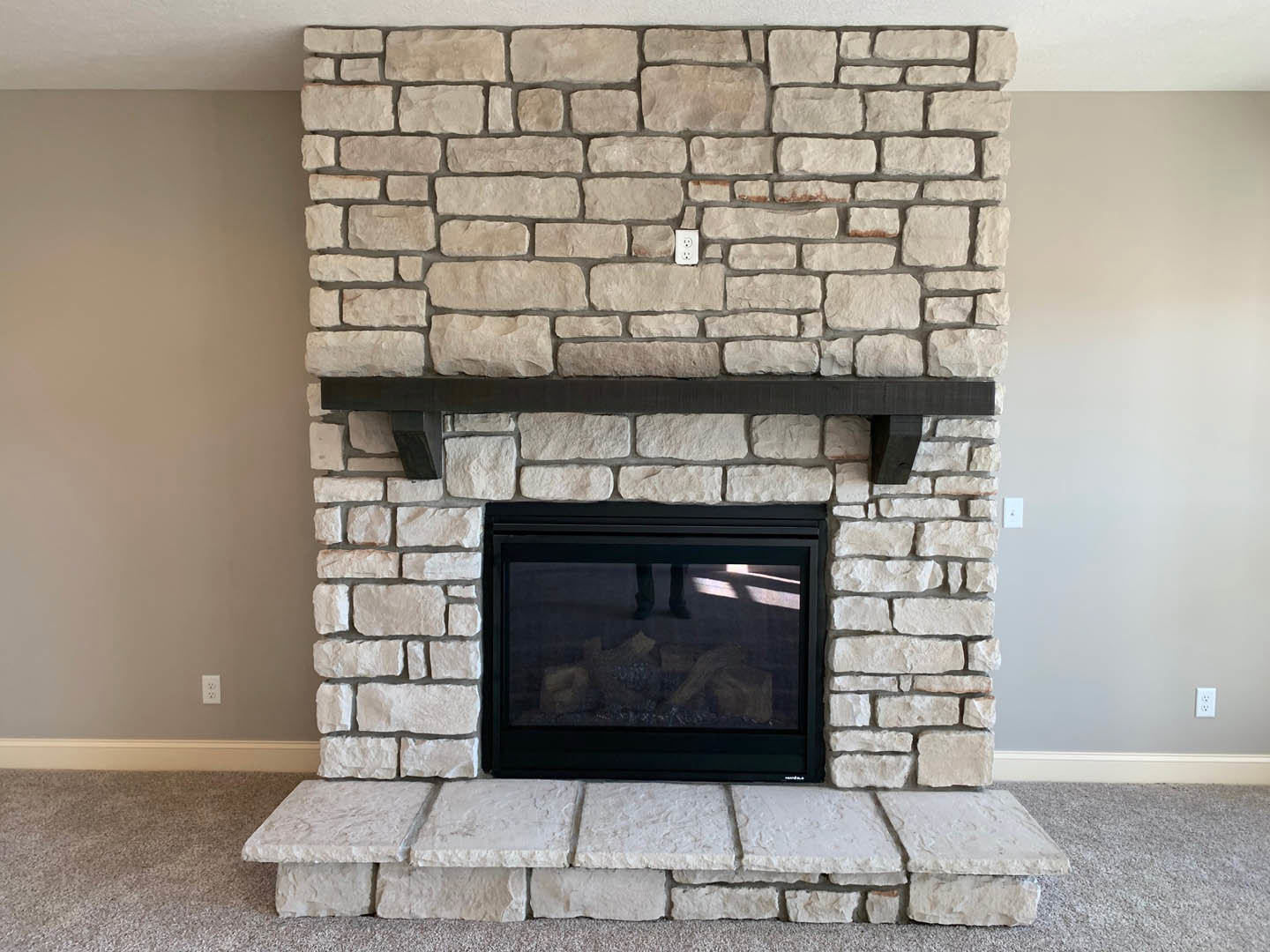 Brick fireplace with glass door and black fire screen, stone hearth on beige carpet, white walls with black trim, wood shelf above masonry oven