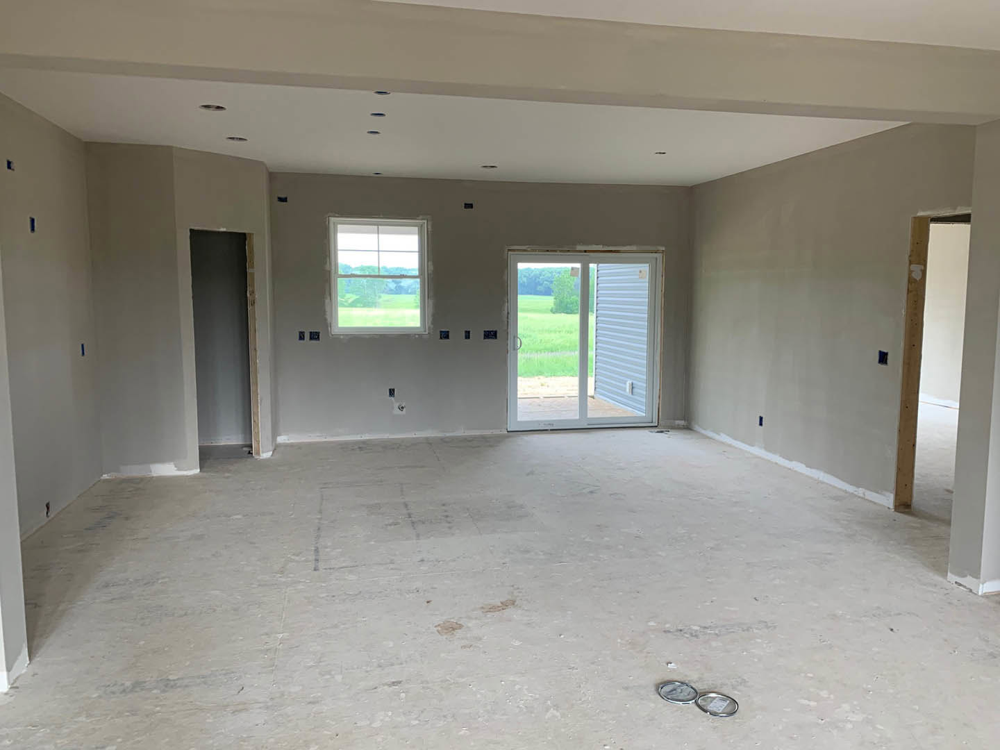 Spacious room featuring a sliding glass door and large windows overlooking a grassy field, concrete flooring, white central fixture, and plaster walls.