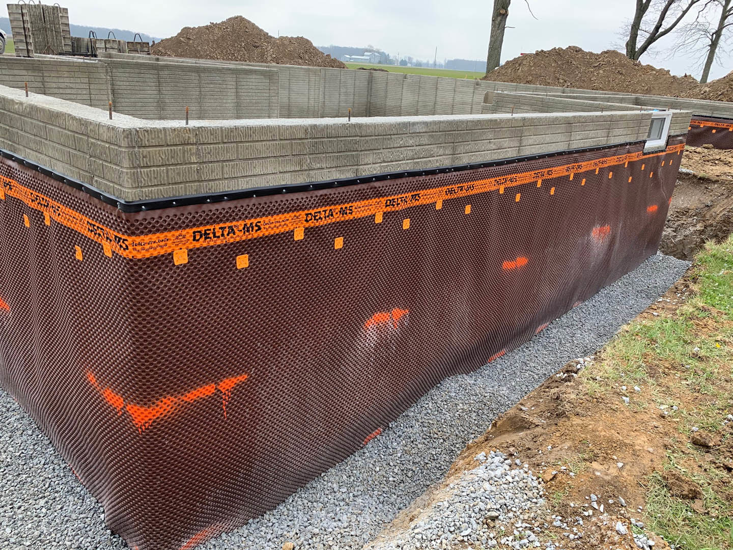 Concrete foundation under construction with gravel base, adjacent to a wooden fence, dirt pile, and partially visible window on nearby building.