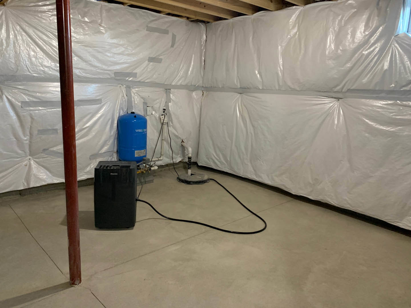 Room with translucent plastic wall coverings, blue cylindrical container with white text, black rectangular object with handle, exposed pipe and hose on unfinished floor.