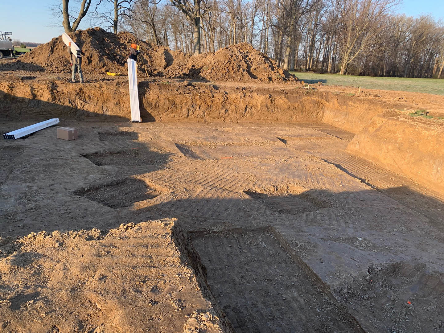 Construction site with large dirt piles, scattered white metal beams, bare trees in background, and a worker carrying lumber