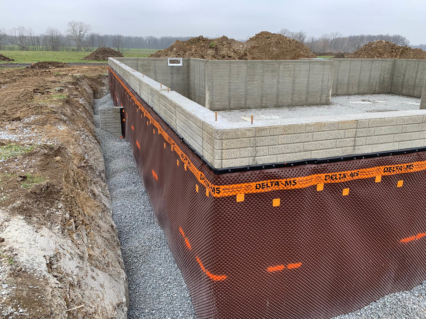 Concrete foundation with brown mesh covering, surrounded by dirt and gravel, adjacent to a partially built wall and hillside under open sky